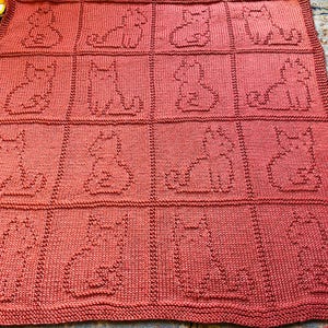 Op de afbeelding: Een koraalkleurige gebreide deken met een herhalend patroon van katten silhouetten. De katten zijn omlijnd in dezelfde koraalkleur als de deken, waardoor een getextureerd, gezellig uiterlijk ontstaat. De deken is rechthoekig en lijkt handgemaakt.