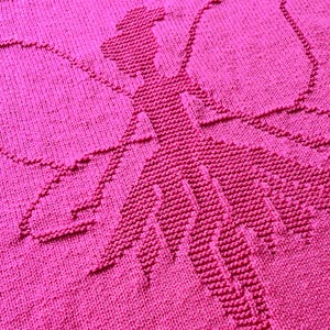 May include: A vibrant hot pink knitted blanket featuring a textured, raised design of a fairy with large wings. The pattern is created using knit and purl stitches, forming a silhouette of a figure with a dress and wings against a flat background.