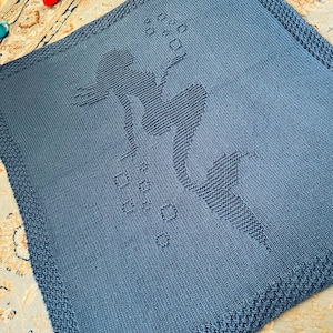 May include: A knitted blue blanket featuring a mermaid design. The mermaid is depicted with flowing hair and bubbles. The blanket has a textured border and is made of soft yarn, perfect for a baby or child.