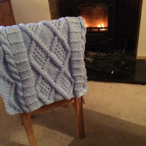 May include: A light blue knitted throw blanket with a cable knit pattern is draped over a wooden chair in front of a fireplace.