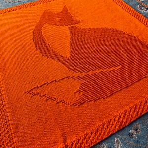 May include: A knitted orange blanket with a cat silhouette design. The blanket is made with a textured stitch and has a fringed border.