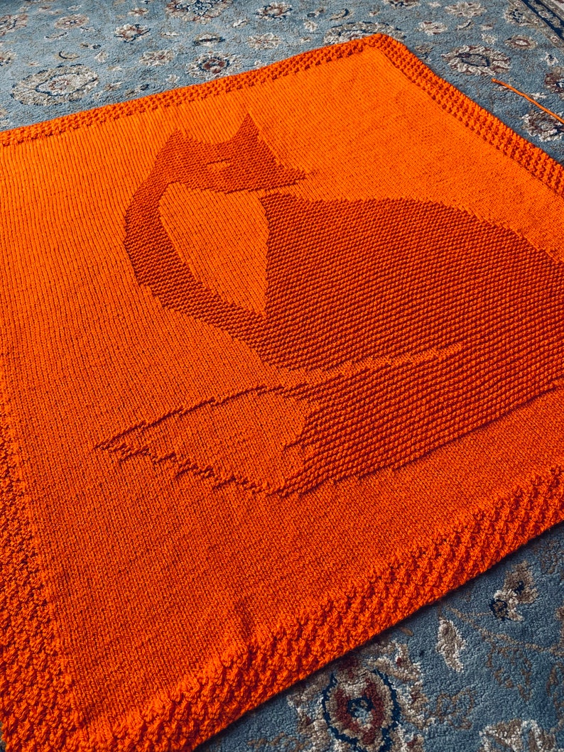 May include: A knitted orange blanket with a cat silhouette design. The blanket is made with a textured stitch and has a fringed border.