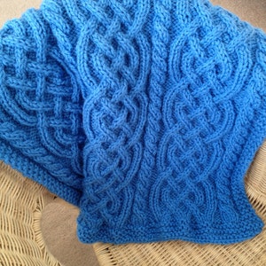 May include: A blue knitted blanket with a cable knit pattern. The blanket is draped over a wicker chair.