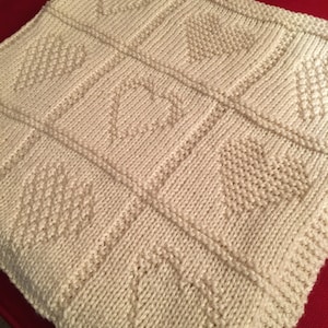 May include: A cream-colored knitted blanket featuring a grid pattern of squares. Each square contains a textured heart design created with purl stitches against a background of stockinette stitches, framed by raised knit borders.