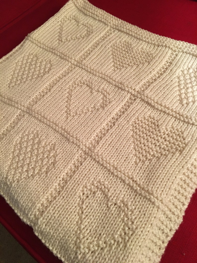 May include: A cream-colored knitted blanket featuring a grid pattern of squares. Each square contains a textured heart design created with purl stitches against a background of stockinette stitches, framed by raised knit borders.