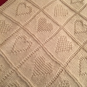 May include: A cream-colored knitted blanket featuring a repeating grid pattern of squares. Each square contains a textured heart motif created with purl stitches against a stockinette background, separated by raised vertical and horizontal borders.