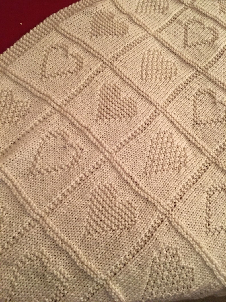 May include: A cream-colored knitted blanket featuring a repeating grid pattern of squares. Each square contains a textured heart motif created with purl stitches against a stockinette background, separated by raised vertical and horizontal borders.