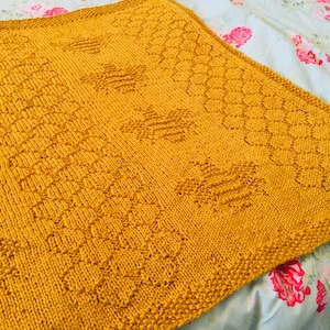 May include: A mustard yellow knitted blanket with a geometric pattern. The blanket is made with a textured yarn and has a soft, cozy feel.