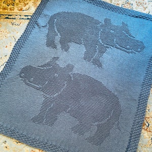 Puede incluir: Una manta de bebé de punto gris con dos hipopótamos bordados. La manta está hecha de un hilo suave y texturizado.