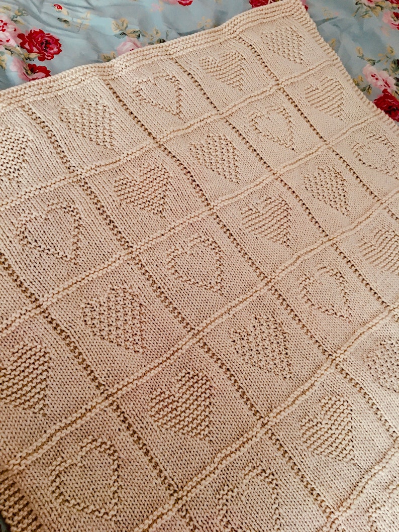 May include: A cream-colored knitted blanket featuring a repeating grid pattern of textured hearts. Each heart is formed by a distinct knit stitch pattern within a square border, creating a uniform and structured design across the entire fabric surface.