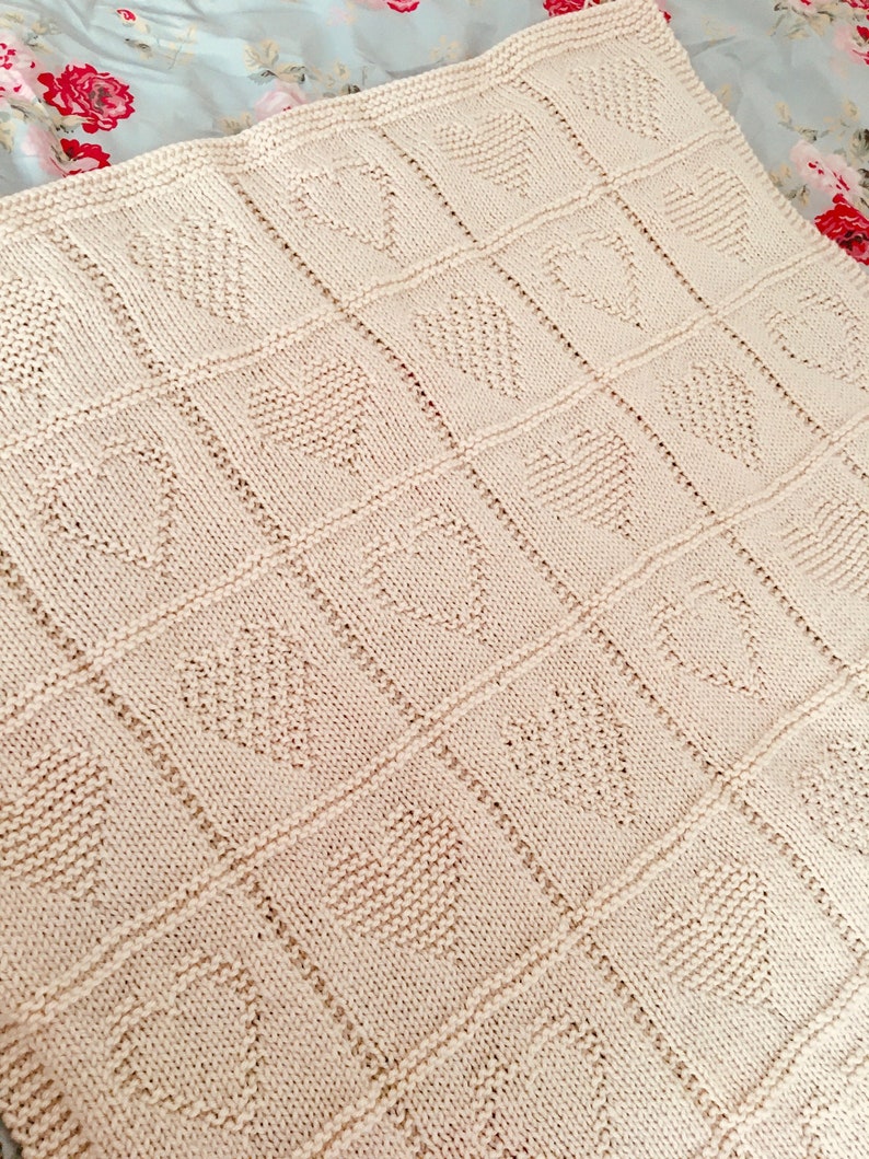 May include: A cream-colored knitted blanket featuring a repeating grid pattern of textured hearts. The blanket is constructed with a garter stitch border and vertical and horizontal lines separating each individual heart square design.