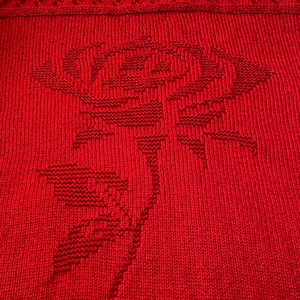 Puede incluir: Una manta de punto roja con un diseño floral. La manta está hecha de un hilo suave y texturizado y presenta una gran rosa con hojas.