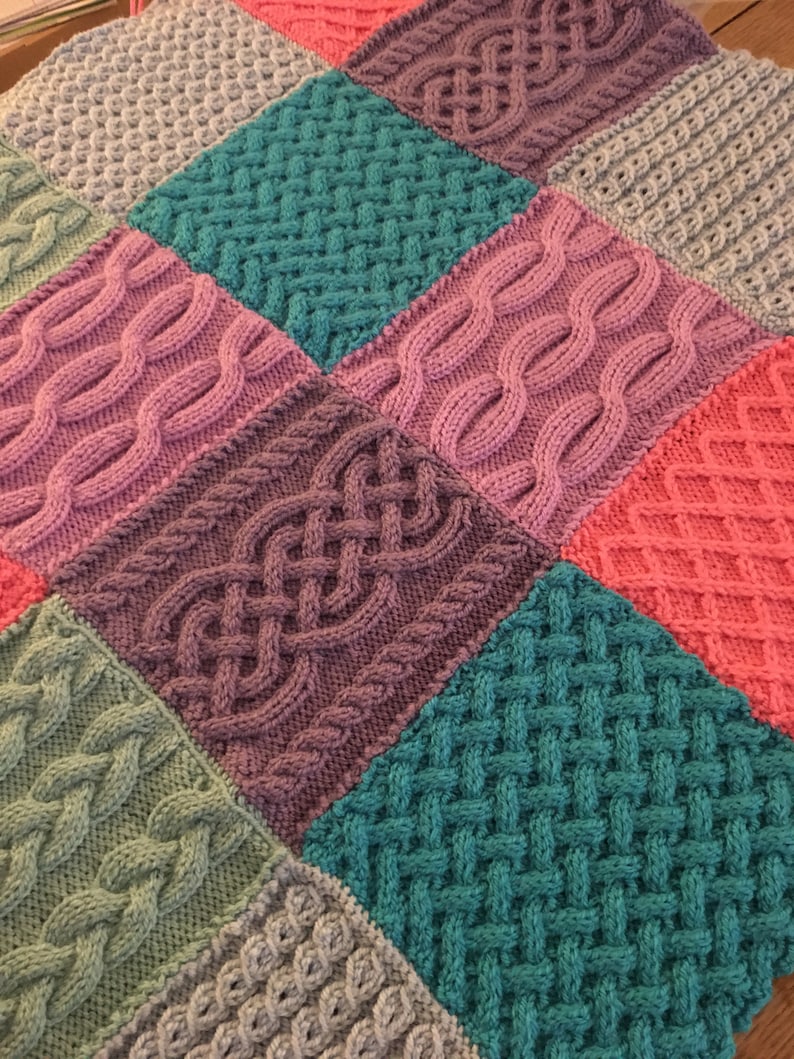 Peut inclure: Une couverture tricot&eacute;e avec un motif patchwork pr&eacute;sentant diff&eacute;rents motifs de tricot en c&acirc;ble dans des tons de rose, violet, turquoise, vert et gris.