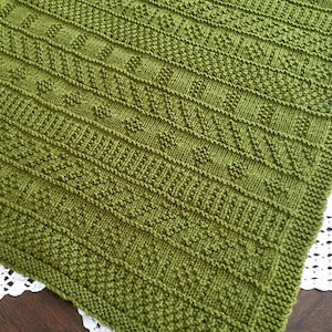 May include: A green knitted blanket with a geometric pattern. The blanket is made with a textured stitch and has a subtle sheen.