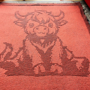 May include: A reddish-orange knitted blanket with a black bull design. The bull's face, horns, and body are clearly defined, creating a striking contrast. The blanket is rectangular and appears handmade. The blanket is likely made of wool or a similar material.