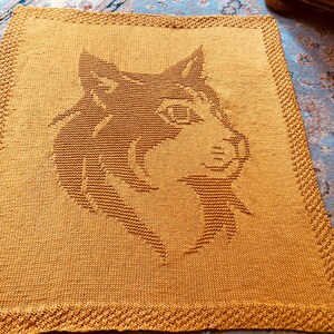 May include: Mustard yellow knitted blanket featuring a detailed wolf head design in a darker shade of brown. The blanket has a textured appearance with a decorative border. A cozy and warm home decor item.