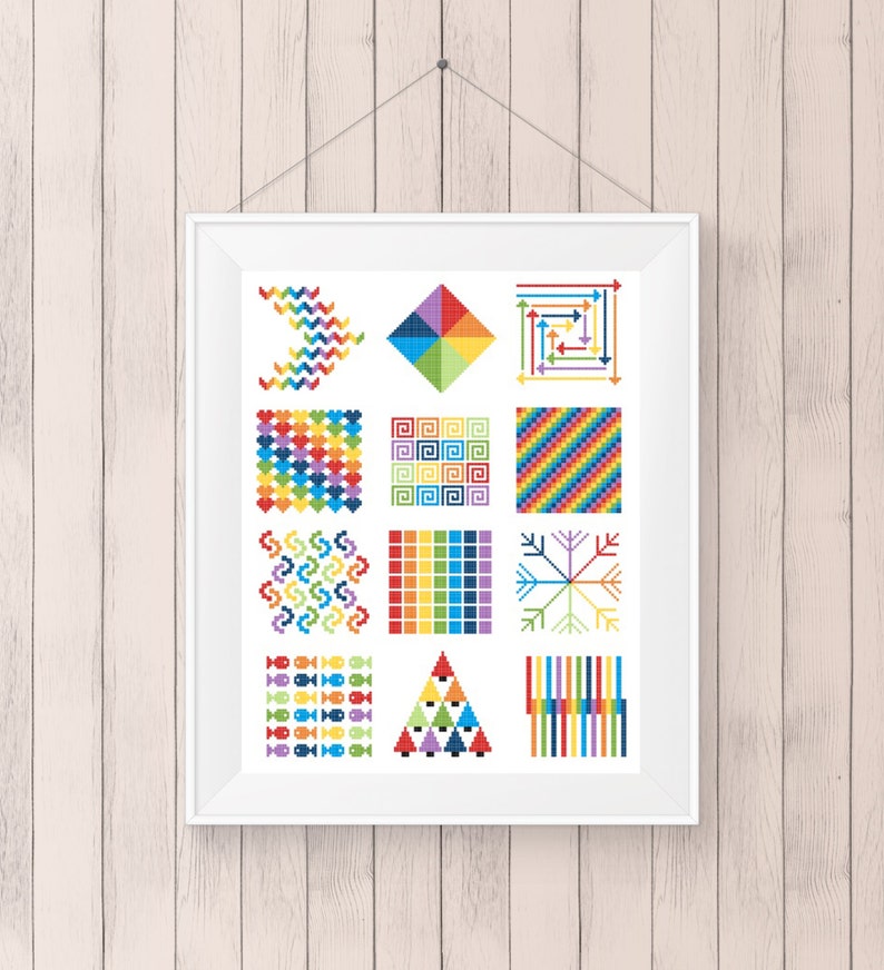 May include: A framed artwork featuring a collection of colorful cross-stitch patterns. The designs include geometric shapes, zigzags, spirals, and fish, all rendered in a vibrant palette of rainbow hues. The artwork is displayed against a light wood-paneled wall.