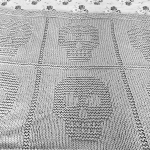 May include: A grey knitted throw blanket with a repeating pattern of skull designs.