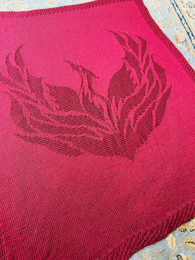 May include: A crimson red knitted blanket with a large, intricate design of stylized flames in the center.
