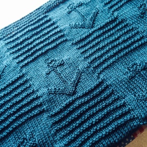 May include: A knitted teal blanket with an anchor pattern. The blanket has a textured design with horizontal ribbed sections and anchor motifs. The colour is a deep, ocean-like teal, creating a cosy and nautical feel.