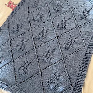 May include: A light blue knitted baby blanket with a repeating pattern of bunny rabbit shapes. The blanket features a diamond grid design with the bunny figures in each diamond. The edges are finished with a ribbed border.