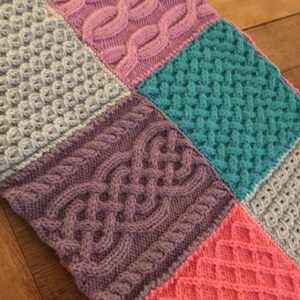 Peut inclure: Une couverture tricot&eacute;e avec un motif patchwork pr&eacute;sentant diff&eacute;rents motifs de tricot en c&acirc;ble dans des tons de rose, violet, gris, turquoise et corail.