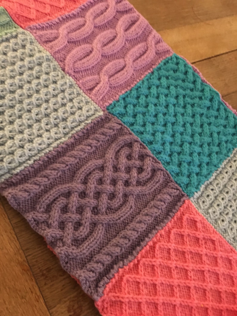 Peut inclure: Une couverture tricot&eacute;e avec un motif patchwork pr&eacute;sentant diff&eacute;rents motifs de tricot en c&acirc;ble dans des tons de rose, violet, gris, turquoise et corail.