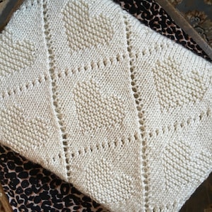 May include: Cream-colored knitted blanket with a heart-shaped design. The blanket features a textured pattern with diagonal lines and small eyelet details. The blanket is folded, revealing the intricate knitwork.