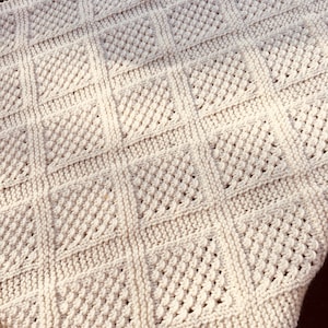 May include: A white knitted blanket with a diamond pattern. The blanket is made with a soft yarn and has a subtle texture.