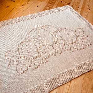 Puede incluir: Una manta de punto rectangular de color beige claro con un diseño central texturizado de tres calabazas rodeadas de hojas. La manta tiene un borde decorativo ancho con un patrón de puntadas en relieve que se repite.