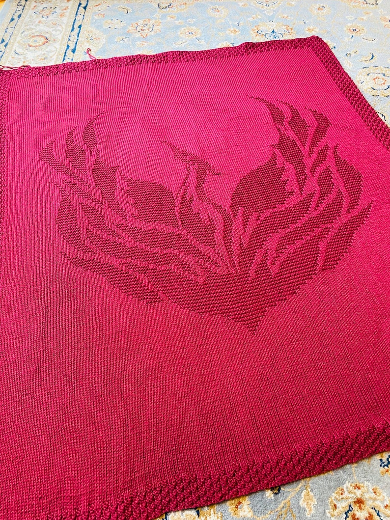 May include: A red knitted blanket with a large, intricate design of a bird with outstretched wings.
