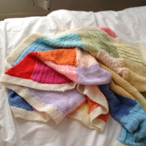 May include: A colorful knitted blanket with squares of different colors, including red, orange, yellow, green, blue, and purple. The blanket is folded and draped over a white bed.