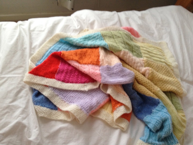 May include: A colorful knitted blanket with squares of different colors, including red, orange, yellow, green, blue, and purple. The blanket is folded and draped over a white bed.