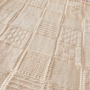 May include: A cream-coloured knitted blanket with a patchwork design. The blanket is made of different knit stitches, creating a textured look.