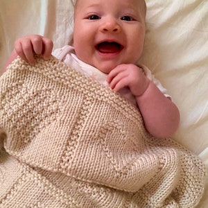 May include: A cream-colored knitted baby blanket featuring a textured geometric pattern with alternating sections of garter stitch and stockinette stitch. The blanket is held up by a smiling infant wearing a white short-sleeved onesie.