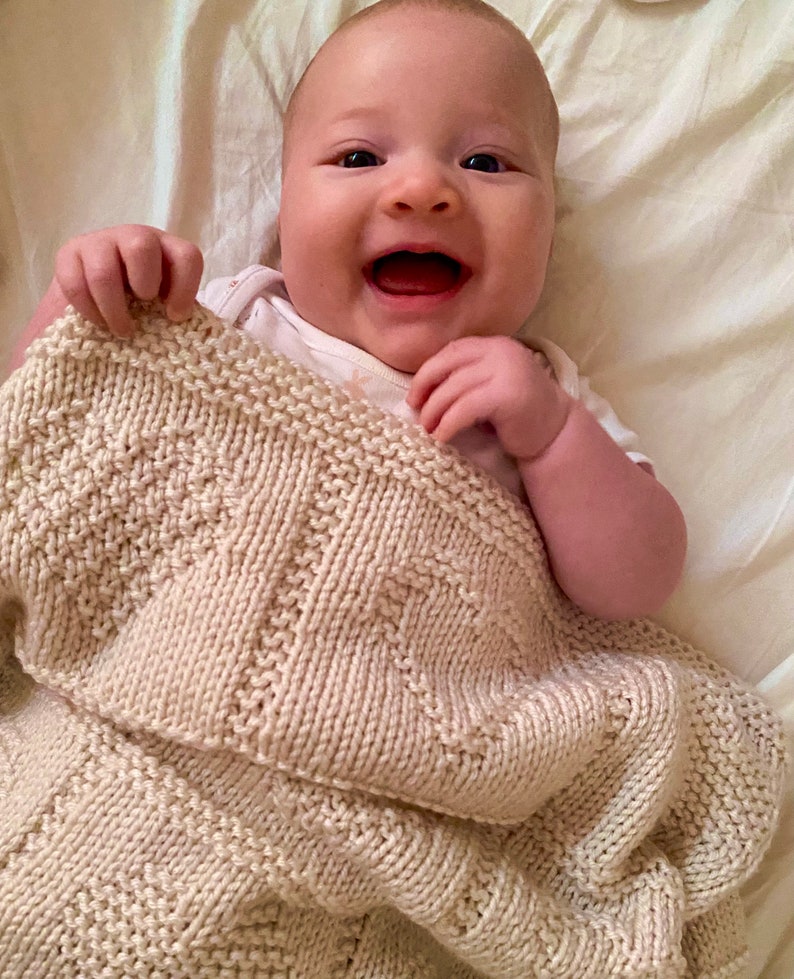 May include: A cream-colored knitted baby blanket featuring a textured geometric pattern with alternating sections of garter stitch and stockinette stitch. The blanket is held up by a smiling infant wearing a white short-sleeved onesie.