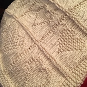 May include: An off-white knitted blanket featuring a repeating pattern of textured hearts. The design is created using a combination of garter stitch and stockinette stitch, with vertical seams separating the panels of the knitted fabric.