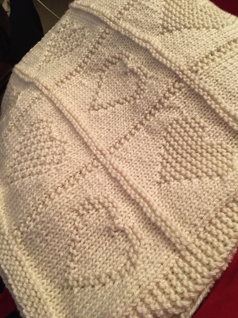 May include: An off-white knitted blanket featuring a repeating pattern of textured hearts. The design is created using a combination of garter stitch and stockinette stitch, with vertical seams separating the panels of the knitted fabric.