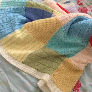 May include: A multi-colored knitted baby blanket with a white border. The blanket is made of different colored squares, including light blue, yellow, green, and pink.