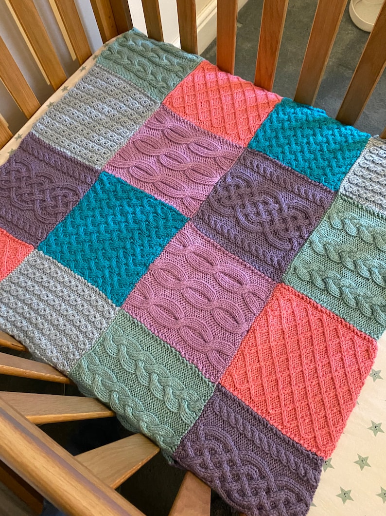 Peut inclure: Une couverture pour b&eacute;b&eacute; en tricot multicolore avec diff&eacute;rents motifs en tricot torsad&eacute; dans des tons de gris, turquoise, rose, vert et violet.