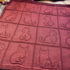 May include: A knitted blanket in a reddish-brown colour, featuring a repeating pattern of cat silhouettes. The blanket is made of a soft, textured yarn, with each cat outlined in a darker shade. The blanket measures approximately 127 cm by 152 cm.
