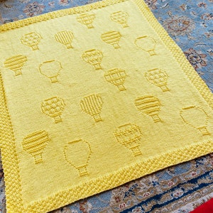 May include: A yellow knitted throw blanket featuring a repeating pattern of hot air balloons. The blanket is made from a soft, textured yarn and has a fringed edge.