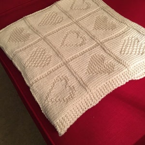 May include: A square, off-white knitted blanket featuring a grid of nine panels. Each panel displays a textured heart motif created with purl stitches against a stockinette background. The edges are finished with a garter stitch border.