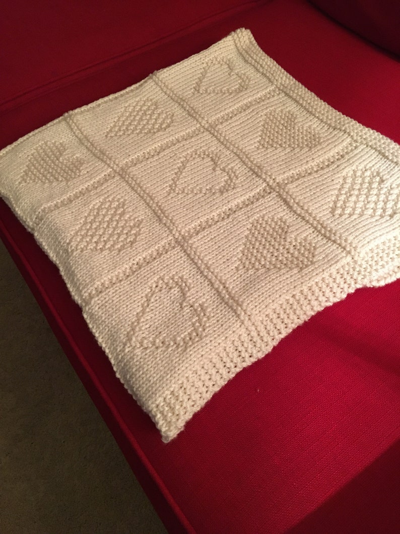 May include: A square, off-white knitted blanket featuring a grid of nine panels. Each panel displays a textured heart motif created with purl stitches against a stockinette background. The edges are finished with a garter stitch border.