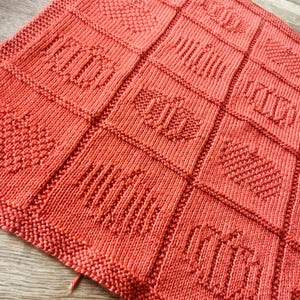 May include: A coral-colored knitted blanket featuring a grid of square panels. Each square displays a distinct textured pattern created with knit and purl stitches, including vertical stripes, diagonal lines, and diamond shapes in a uniform color.
