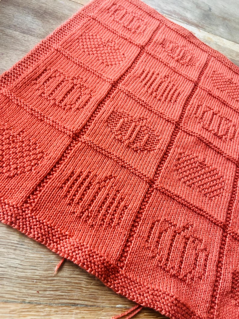 May include: A coral-colored knitted blanket featuring a grid of square panels. Each square displays a distinct textured pattern created with knit and purl stitches, including vertical stripes, diagonal lines, and diamond shapes in a uniform color.