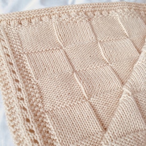 May include: Cream-coloured knitted blanket with a textured, chequered pattern. The blanket features a decorative border with a delicate, openwork design. The soft, neutral colour and detailed stitching suggest a handmade item, suitable for home decor.