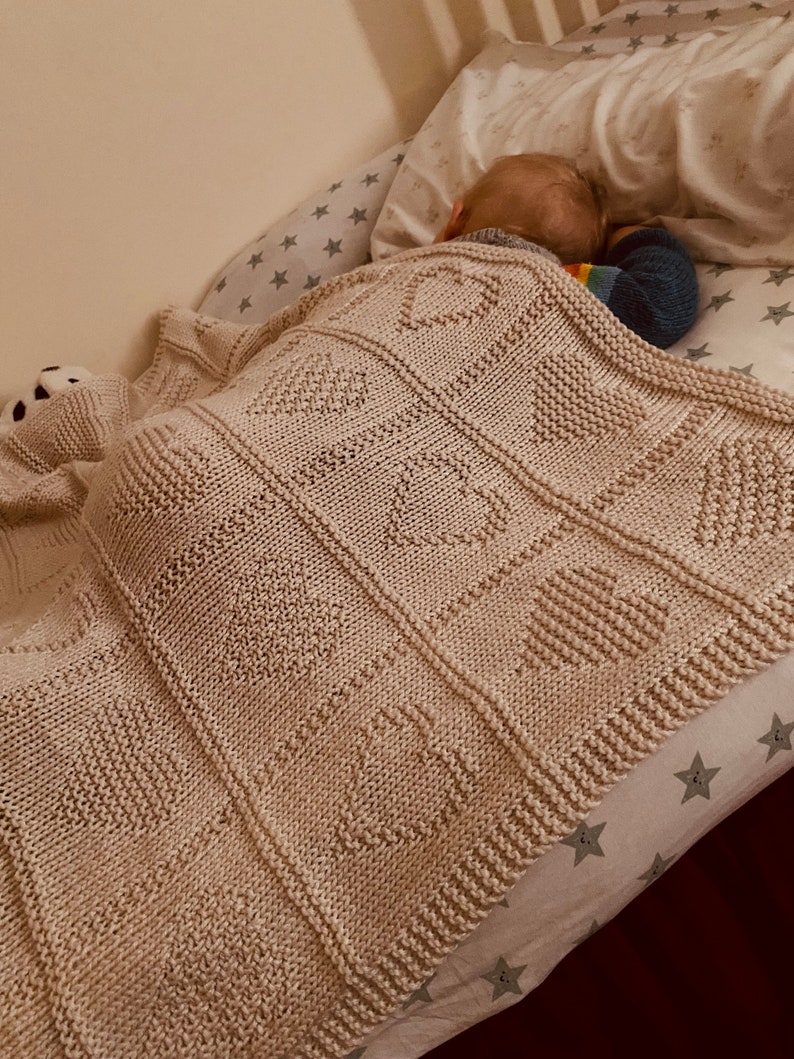 May include: A cream-colored knitted blanket featuring a repeating grid pattern of textured hearts. The blanket is constructed with thick yarn and displays a consistent stitch design throughout each square section, creating a soft and tactile surface.