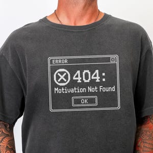 May include: A charcoal grey short-sleeve t-shirt featuring a white pixelated computer error message graphic. The text reads ERROR, 404: Motivation Not Found, and OK, enclosed in a rectangular box with an X icon inside a circle.