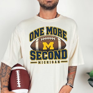 Może przedstawiać: Kremowy t-shirt z krótkim rękawem z grafiką brązowej piłki do futbolu amerykańskiego i żółtą literą M. Napis brzmi ONE MORE SECOND MICHIGAN ciemnoniebieskimi i żółtymi literami. Koszulka jest noszona z brązową piłką trzymaną w jednej dłoni.